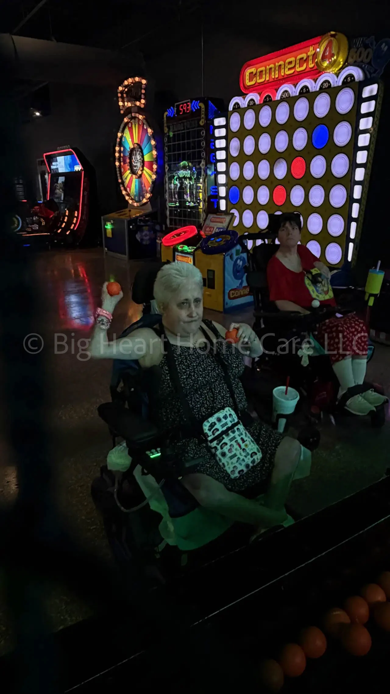 Two Big Heart Group Home residents in power wheelchairs participating in an arcade ball toss game at ELEV8 Fun Tampa, smiling and engaged in the activity.